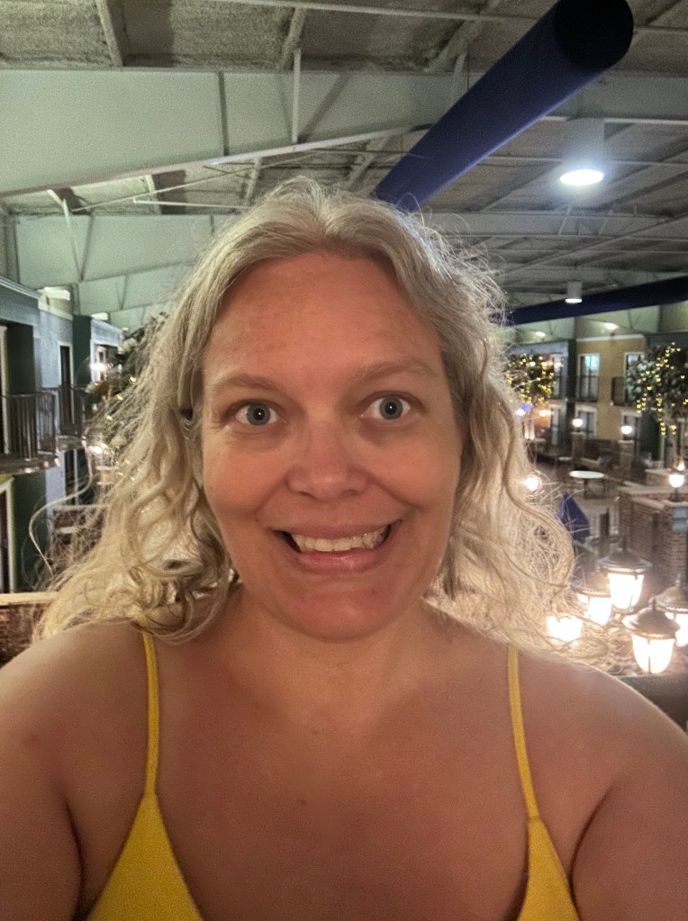 Amanda Freese smiles in a well-lit indoor setting with string lights and balconies in the background. She has shoulder length, wavy, blonde hair and is wearing a yellow spaghetti strap dress. The atmosphere appears warm and festive.