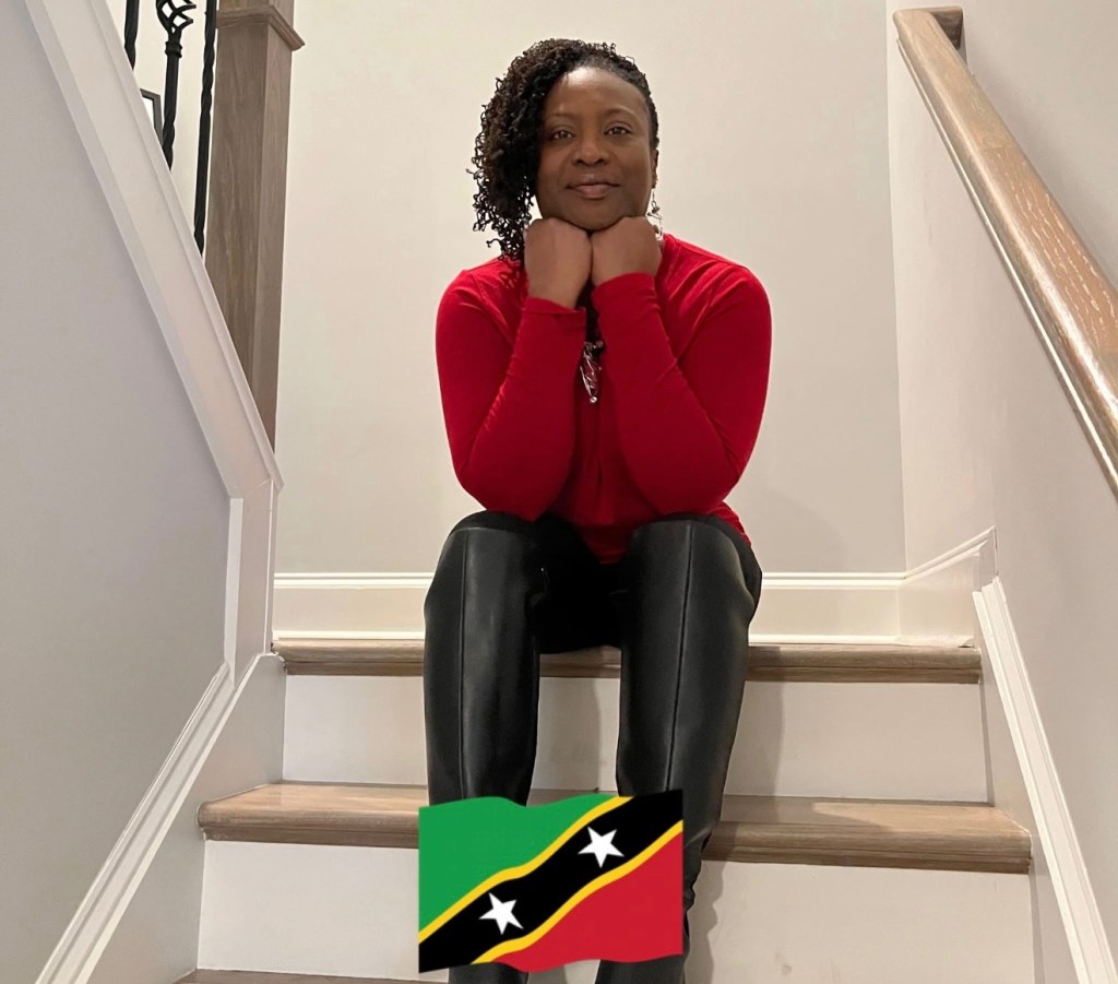 Image of Hatasha sitting confidently on a staircase, wearing a bold red top and sleek black pants. She rests her chin on her hands with calm strength, her expression serene and self-assured. Soft lighting highlights her presence, while the vibrant Saint Kitts and Nevis flag at her feet adds a touch of pride and heritage to the scene.