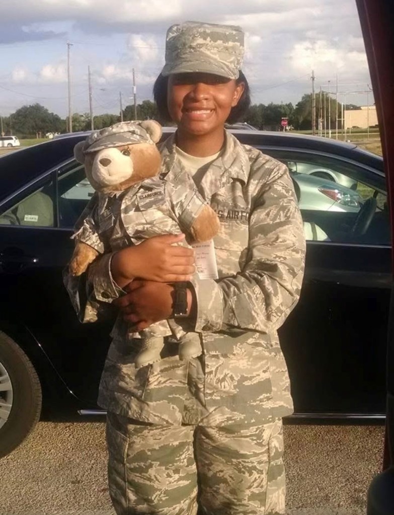 Image of Sisterhood co-founder, Vickie, in her U.S. Air Force military uniform standing outdoors, smiling warmly as she holds a teddy bear dressed in matching camouflage. Parked cars and an open sky frame the scene. 