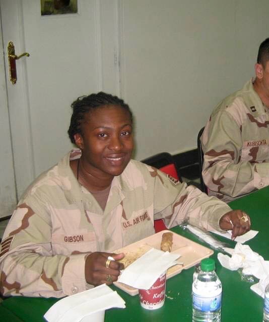 Image of Sisterhood member, Hatasha, in her U.S. Air Force desert camouflage uniform sitting at a table, smiling while holding a fork over a food tray. The uniform’s name tag reads “GIBSON.” A cup, water bottle, and napkins are on the green table. Another service member is partially visible beside her.