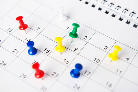 Image of a close-up of a paper calendar page with numbered dates, marked with colorful push pins in red, blue, green, and yellow placed on different days.