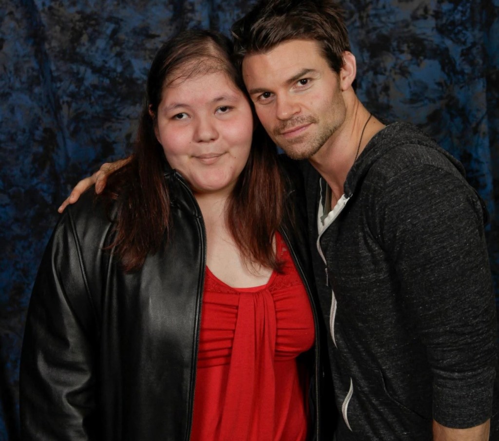 Image of Liliana posing for a photo with a dark-haired man at a meet-and-greet event. Liliana, wearing a red top and black jacket, stands beside the man, who has his arm around her shoulder and is wearing a dark hoodie. Both face the camera and smile slightly, standing in front of a blue patterned backdrop. The man is best known for his role as Elijah in the series Vampire Diaries.