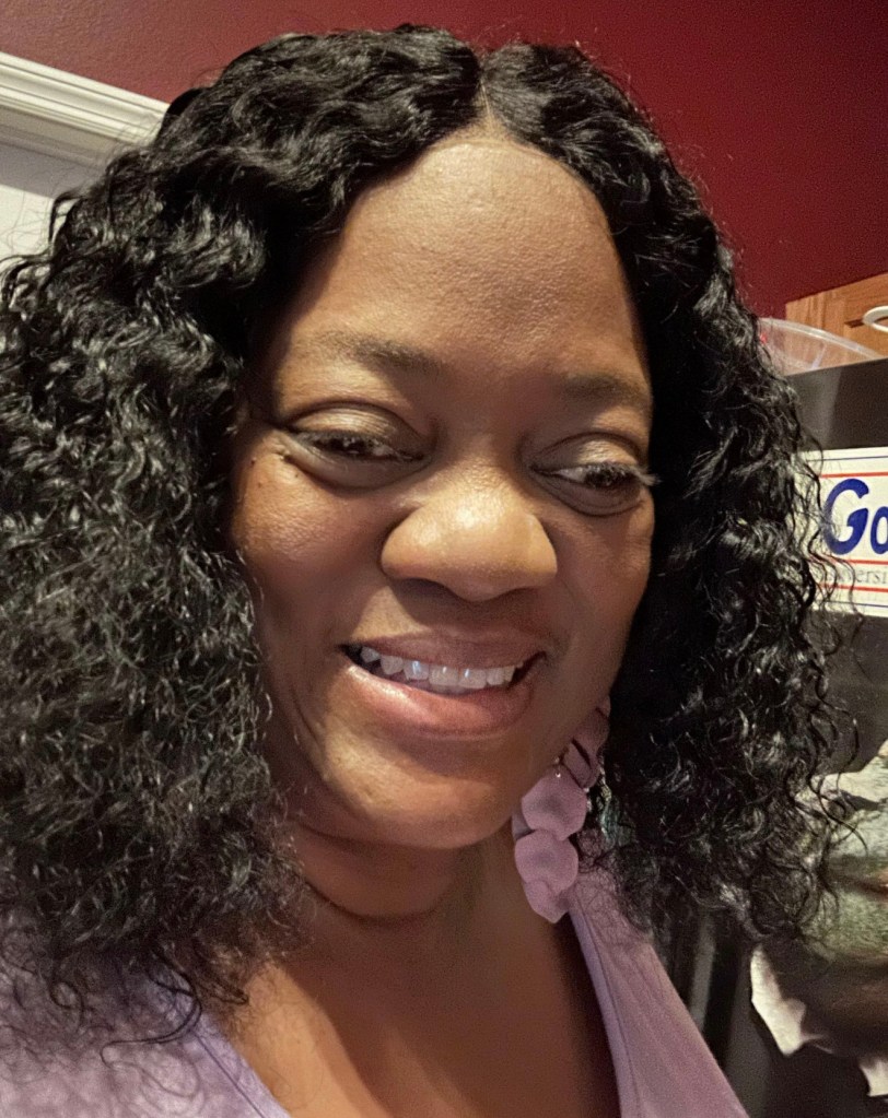 A close-up of a smiling Nanette with shoulder-length, curly black hair. She is indoors, wearing a light purple top, with a warm-toned wall and part of a kitchen visible in the background.