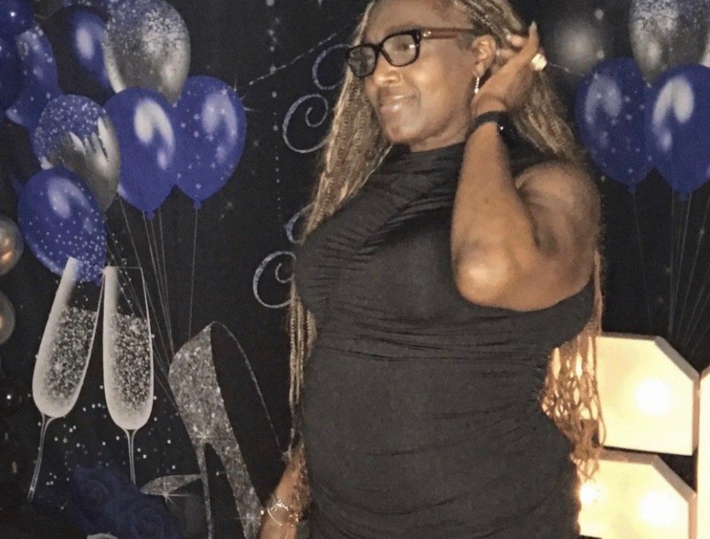 Javiya, this month’s Sister Spotlight winner, stands smiling at a celebration event. She is wearing a black dress and glasses, with long braided hair, and is surrounded by blue and silver balloons and festive decorations in the background.