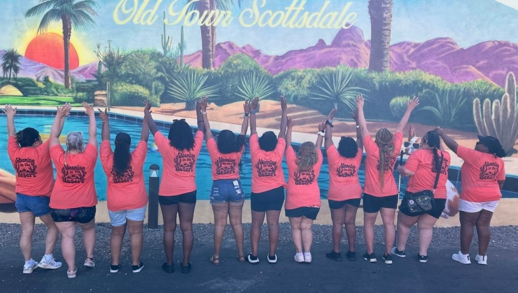 A group of 11 people stand with their backs to the camera in front of a colorful “Old Town Scottsdale” mural featuring a desert landscape, mountains, palm trees, cacti, and a sunset. They are wearing matching bright coral T-shirts that read “Blooming in the Desert” on the back and have their arms raised together in celebration.

